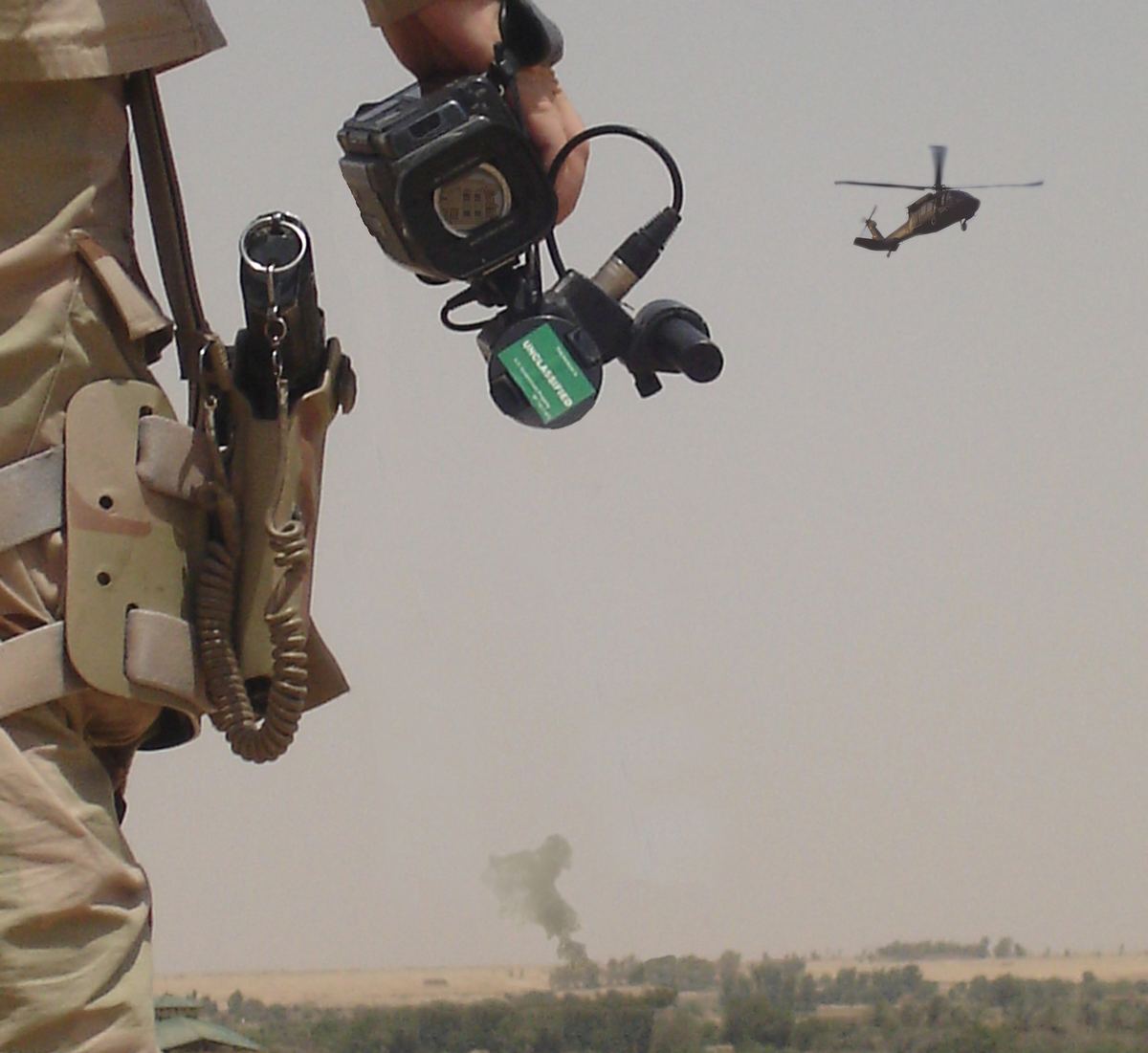 A National Guard soldier stands with a video camera at his side while a Black Hawk helicopter crosses the sky above palm groves and a distant column of smoke in Iraq.